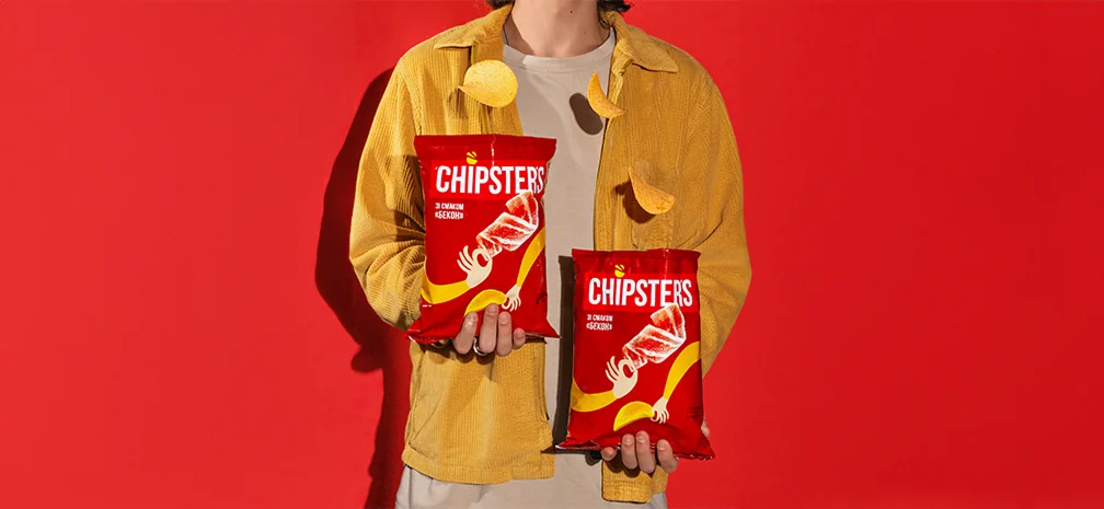 Чоловік тримає два пакети Chipster’s на червоному фоні, чипси в повітрі